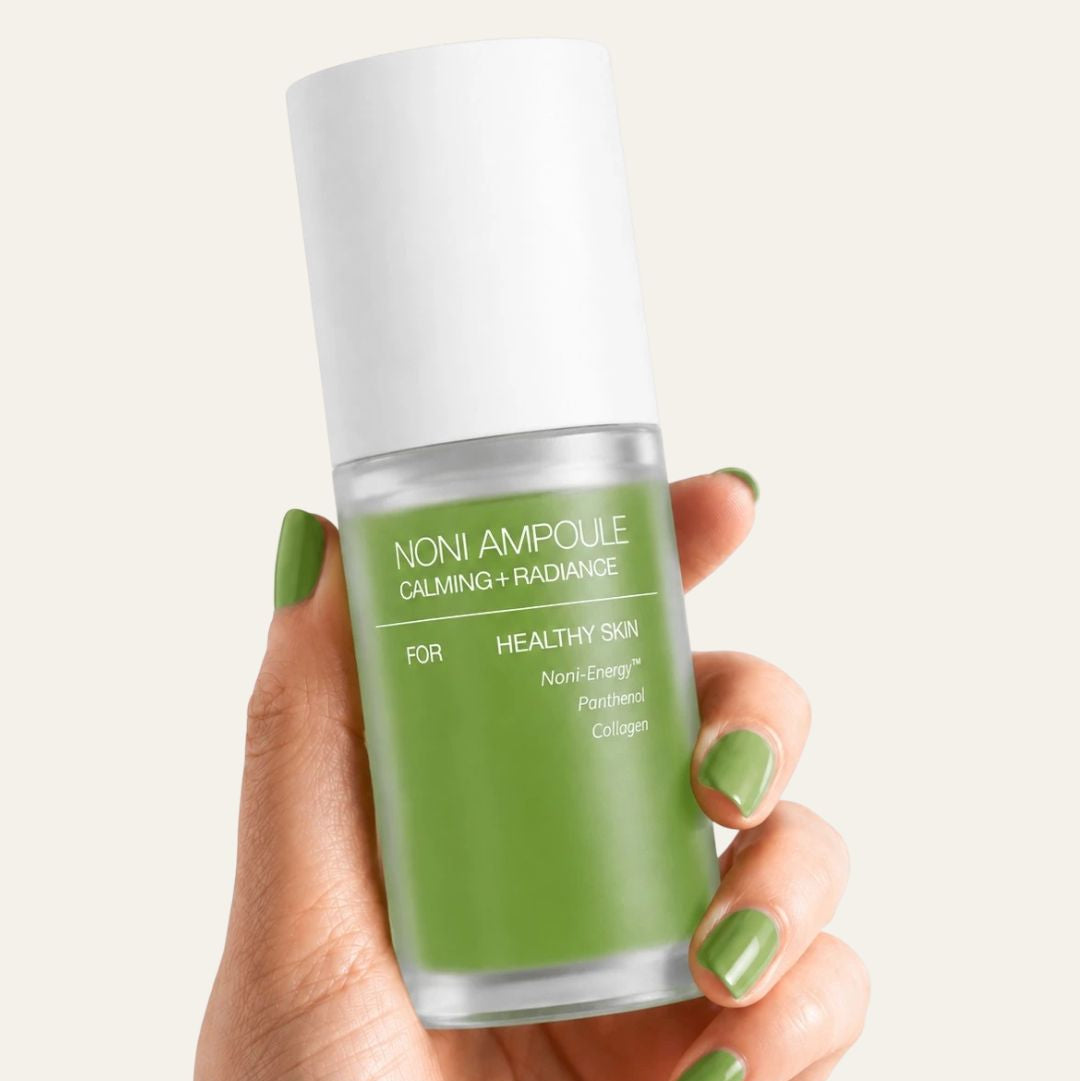 Hand holding a green skincare bottle labeled 'Noni Ampoule' against a white background